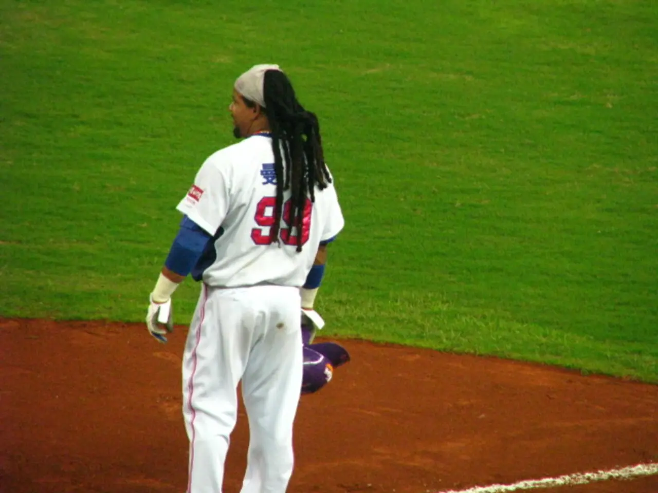 In the image there is a man in white jersey and pants standing on the play field and in the back...