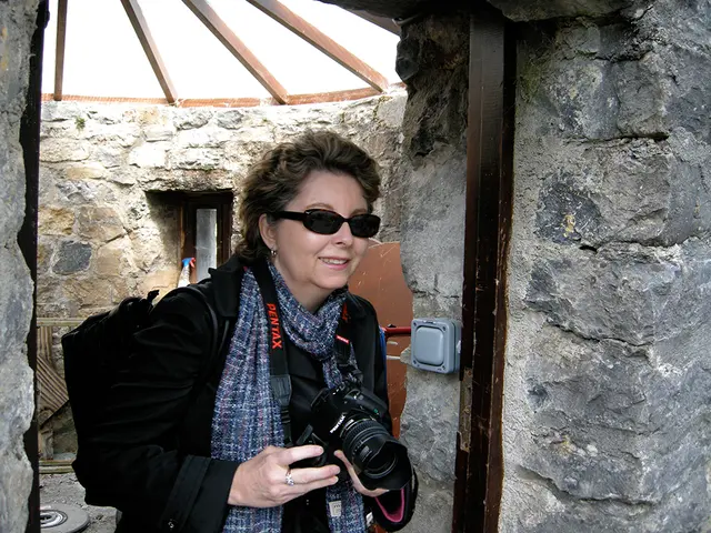 This picture shows a woman standing and holding a camera in her hand we see a sunglasses on her...