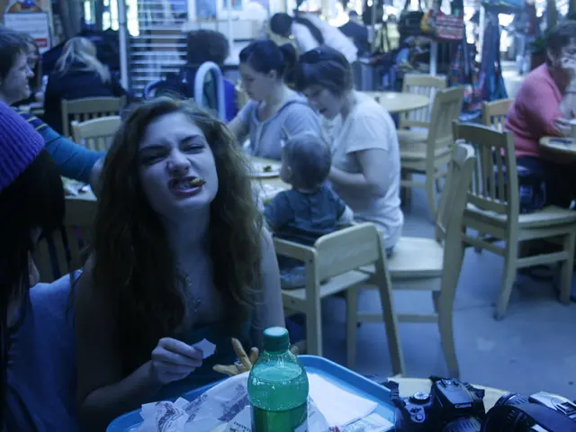 a person is sitting and having food. in front of her there is a bottle and a camera. behind her...